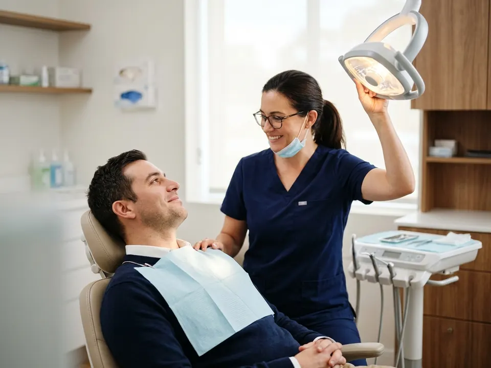 A dentist adjusting a bright clinic light while speaking reassuringly to a patient in a dental chair.